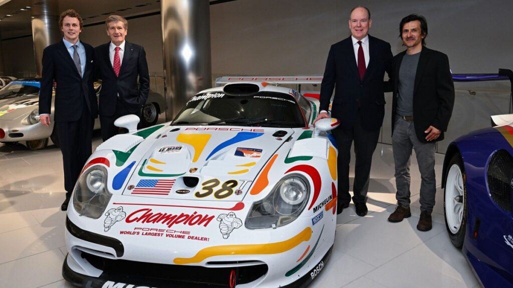 VIPs like Ferdinand Porsche, Stéphane Ortelli, and Thierry Bousten gather around a Porsche racing model. 